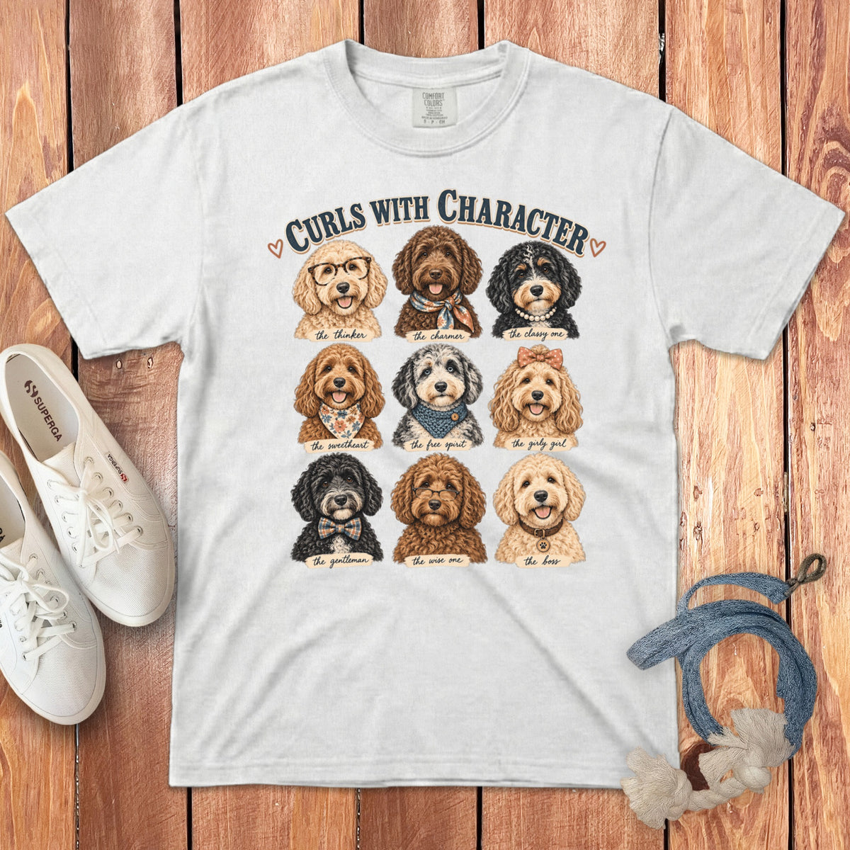 A white crewneck t-shirt featuring a graphic of nine different doodle dogs with text reading Curls With Character, laid flat on a wooden surface alongside white sneakers and a blue rope toy.