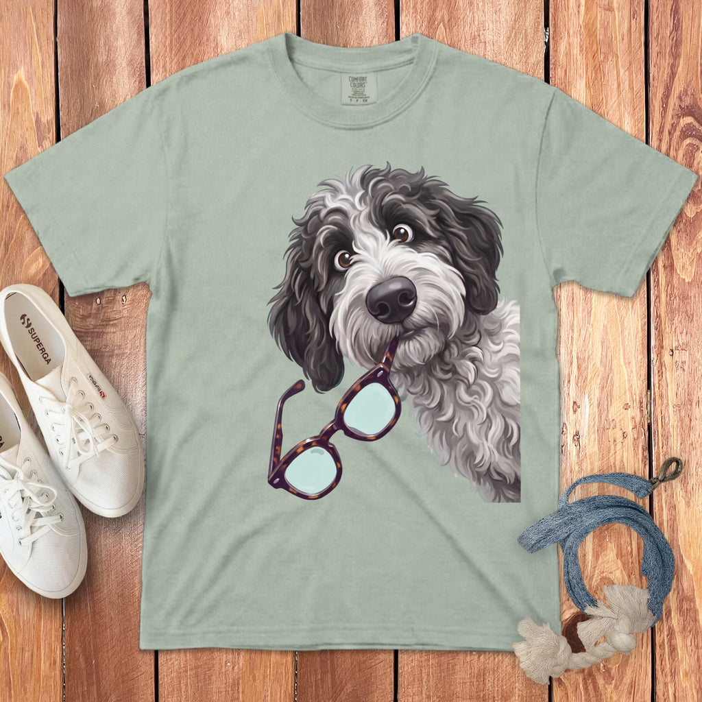 Sage green t-shirt featuring a cartoon dog holding glasses in its mouth, laid on a wooden background.