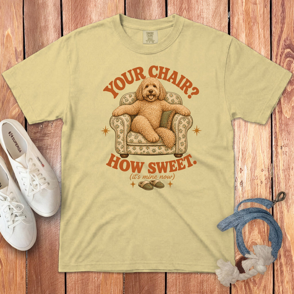 A yellow t-shirt with a graphic of a dog sitting in an armchair, surrounded by text and accessories on a wooden background.