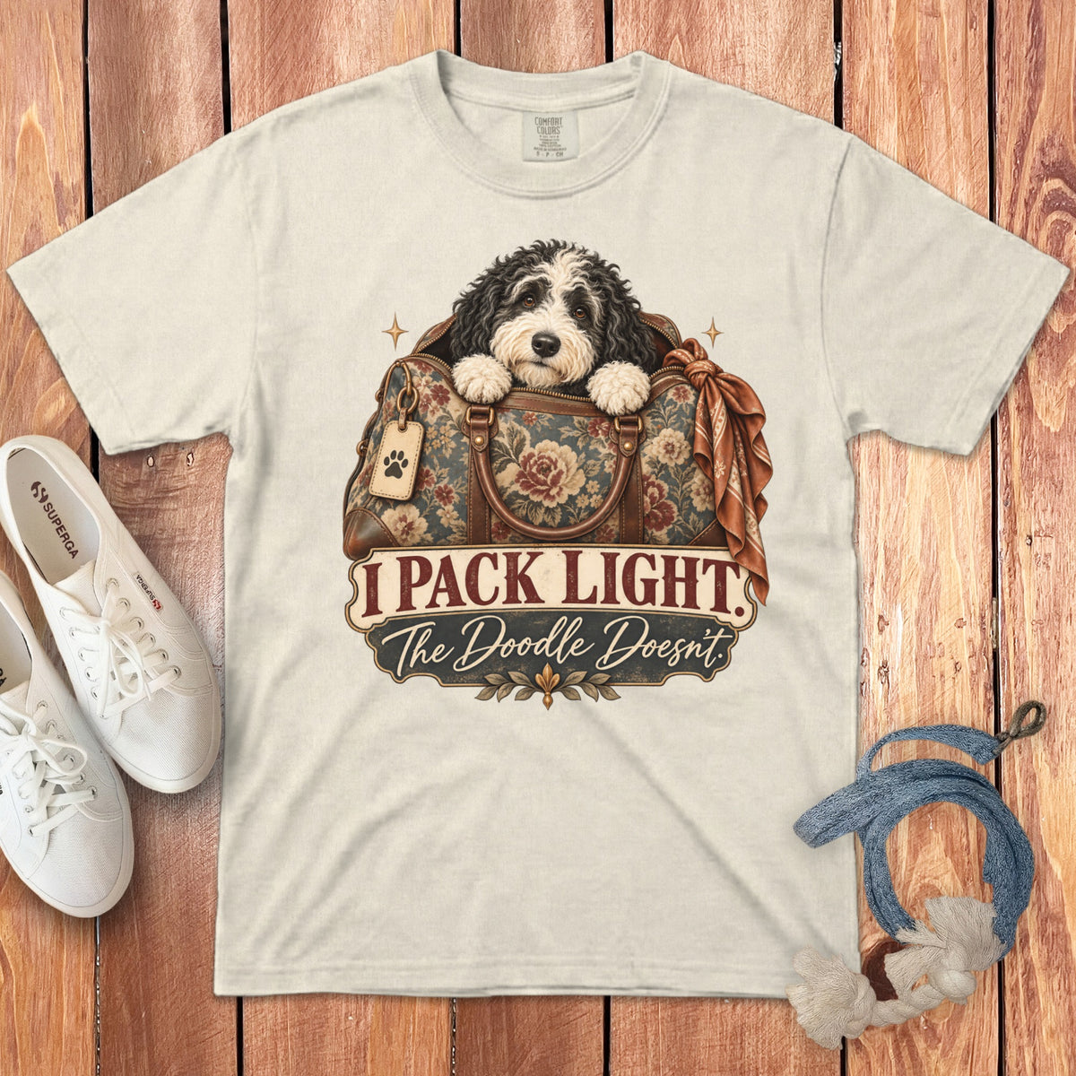 Beige t-shirt featuring a doodle dog inside a floral travel bag with text reading I Pack Light The Doodle Doesnt.