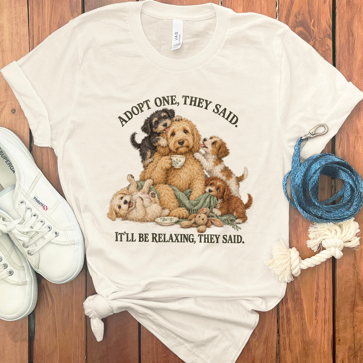 Cream t-shirt with illustration of several goldendoodle puppies surrounding an adult goldendoodle holding a teacup. Text reads "ADOPT ONE, THEY SAID. IT'LL BE RELAXING, THEY SAID."
