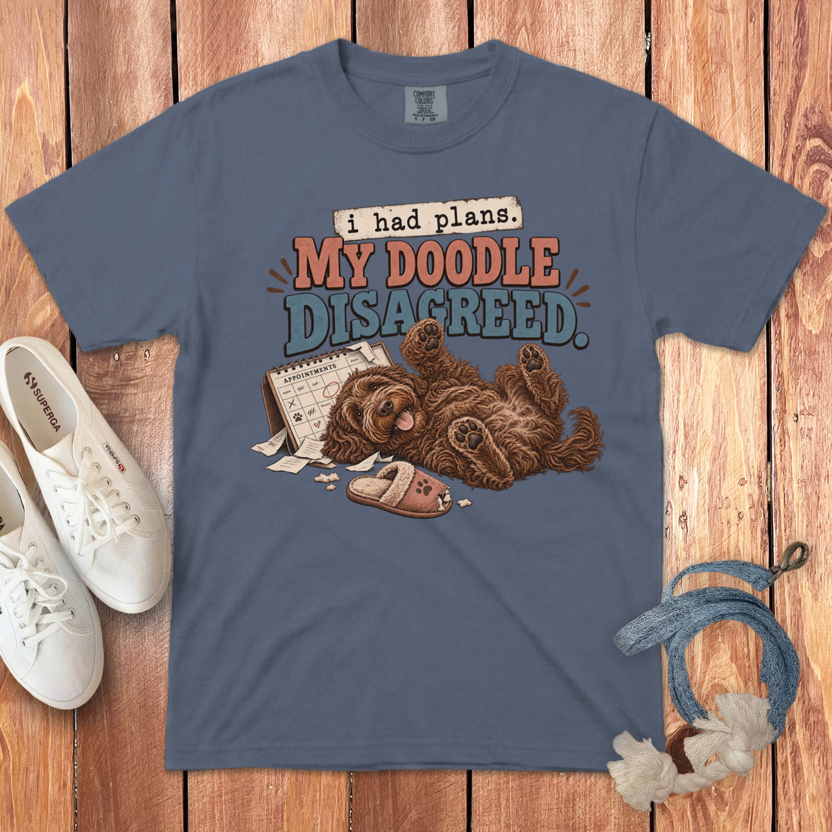 A slate blue t-shirt featuring a playful doodle dog lying on its back with a calendar and chewed slipper.