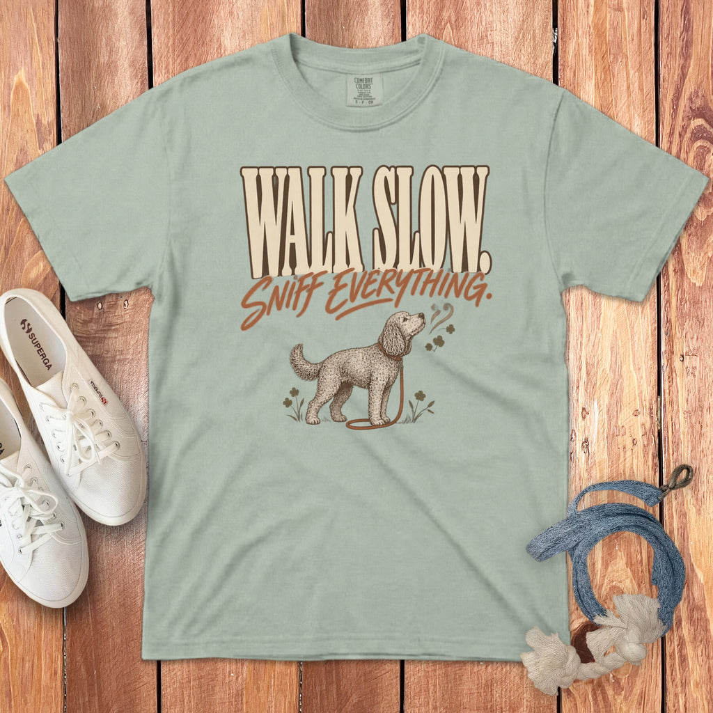 Sage green t-shirt featuring the text "WALK SLOW SNIFF EVERYTHING" and a dog illustration, laid on wood.