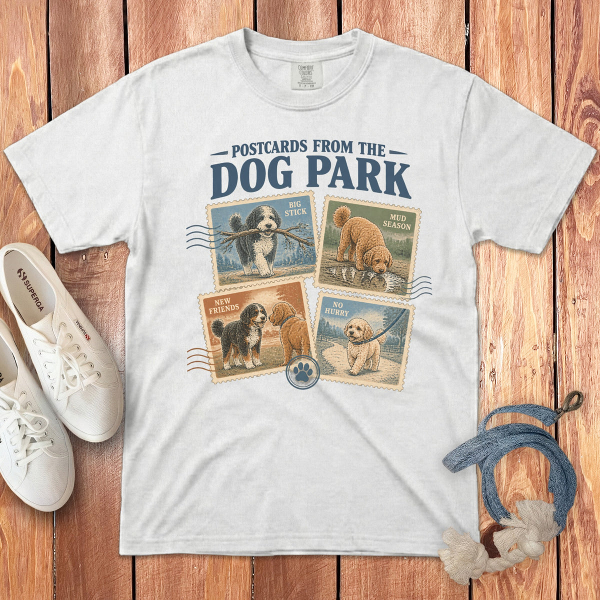 A white t-shirt featuring a Postcards from the Dog Park graphic design with four stamp-style dog illustrations.