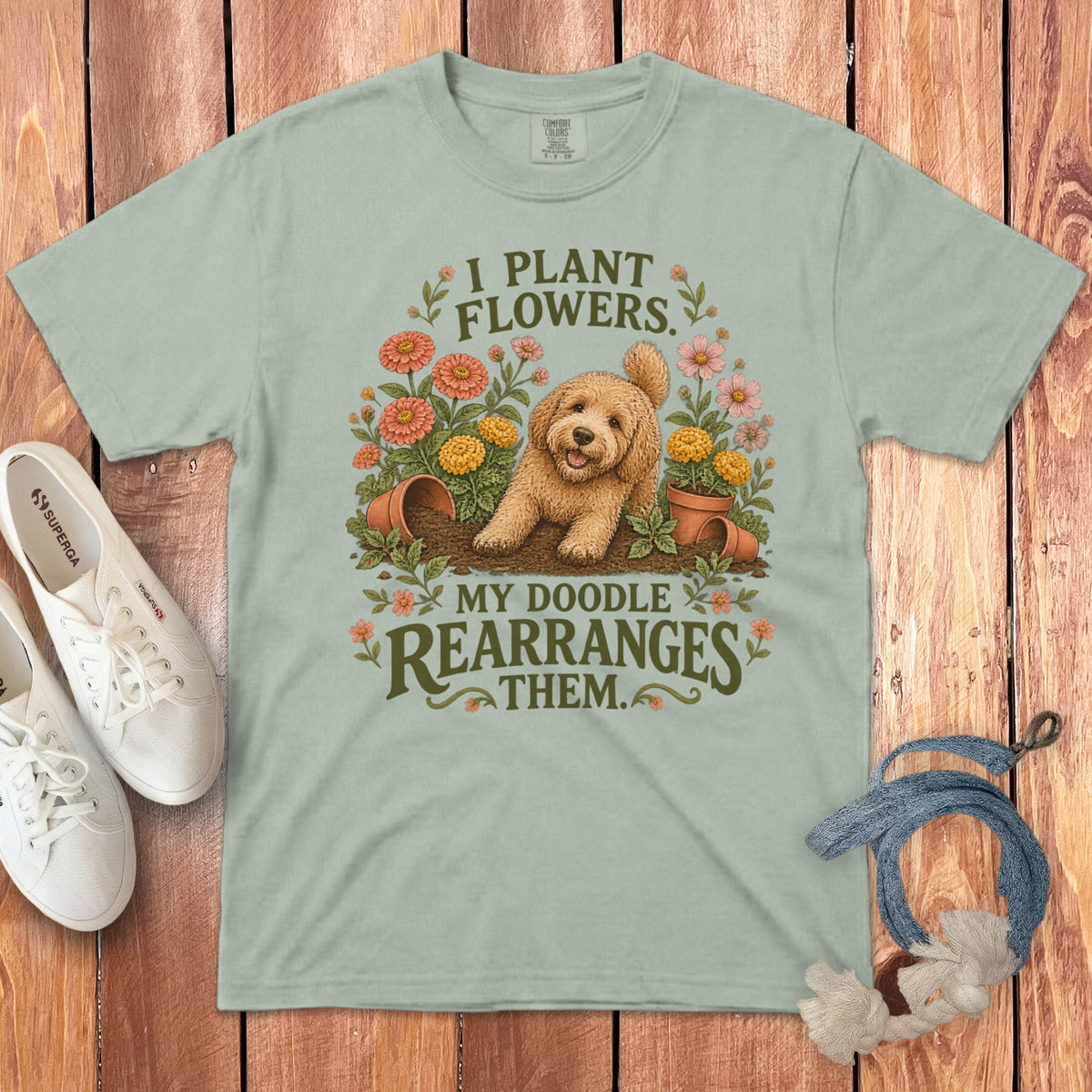Sage green t-shirt featuring a goldendoodle graphic and text reading I plant flowers my doodle rearranges them.