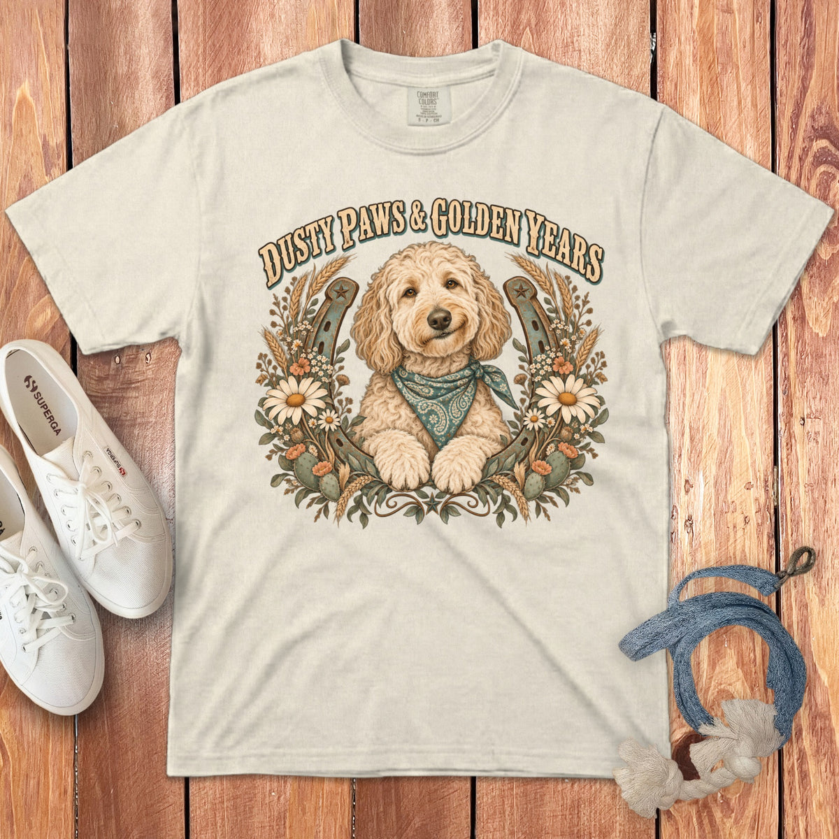 A cream-colored t-shirt with a golden doodle graphic, horseshoe, flowers, and Dusty Paws and Golden Years text.