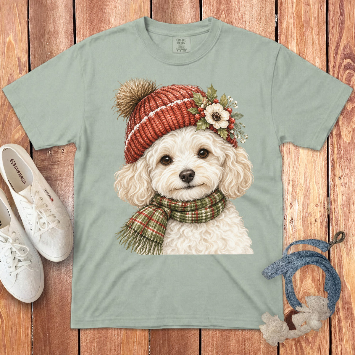 Light green t-shirt with watercolor illustration of a white poodle wearing a red knit hat and scarf.