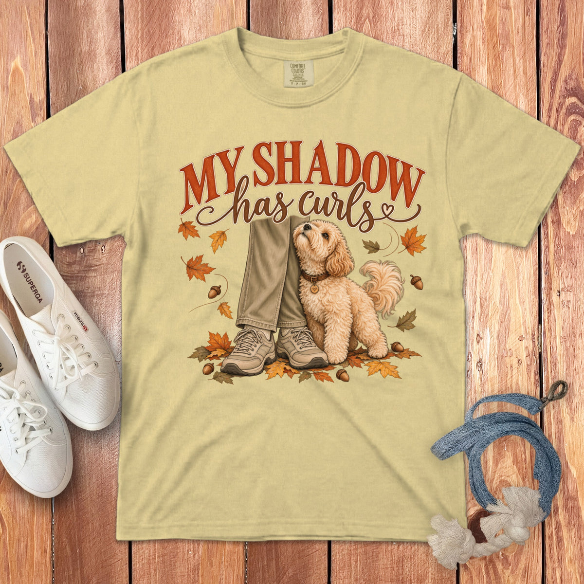 A light yellow t-shirt features a graphic of a small curly dog beside legs, surrounded by autumn leaves and acorns, with the text My shadow has curls.