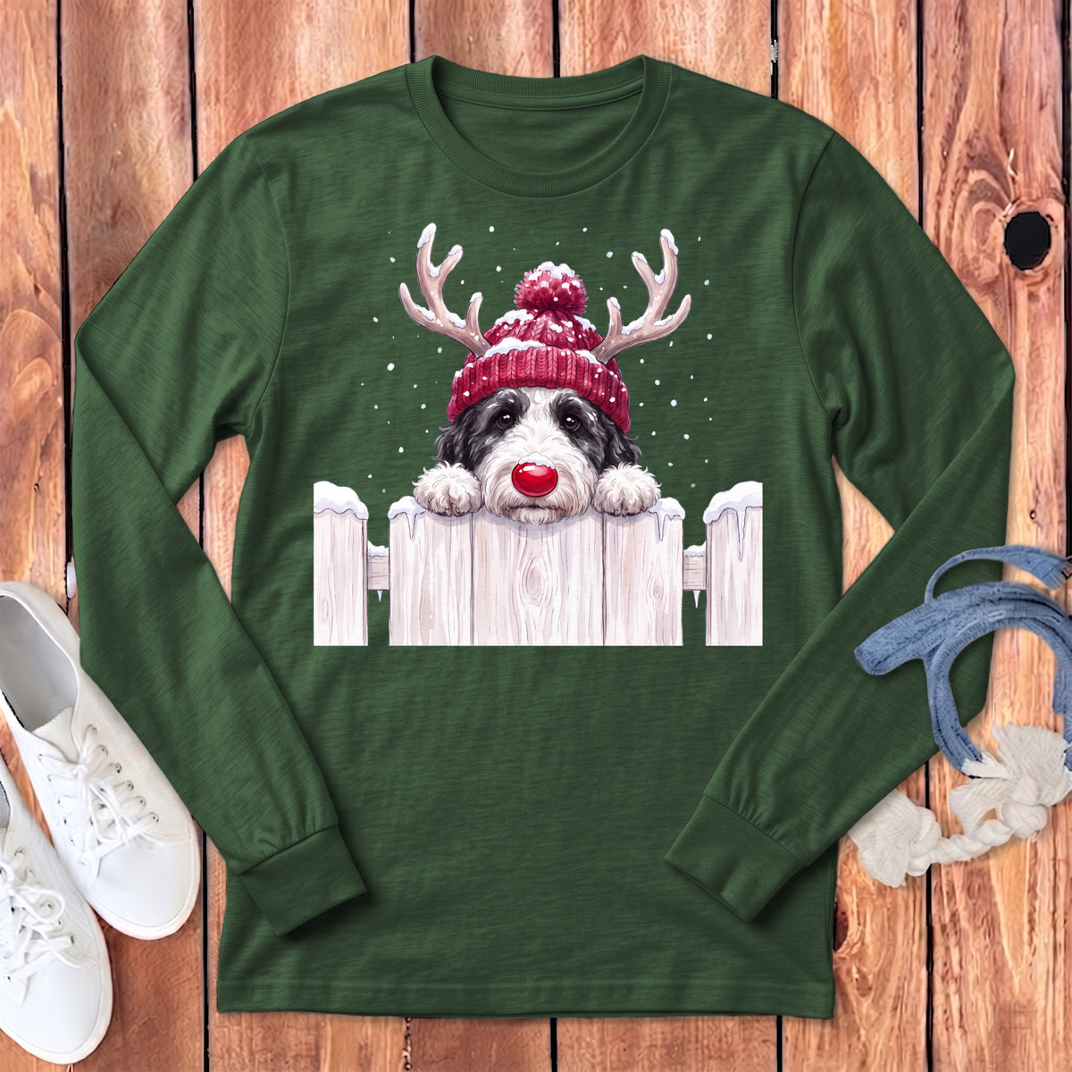 Dog in reindeer hat and red nose peeking over snowy fence on a green shirt.