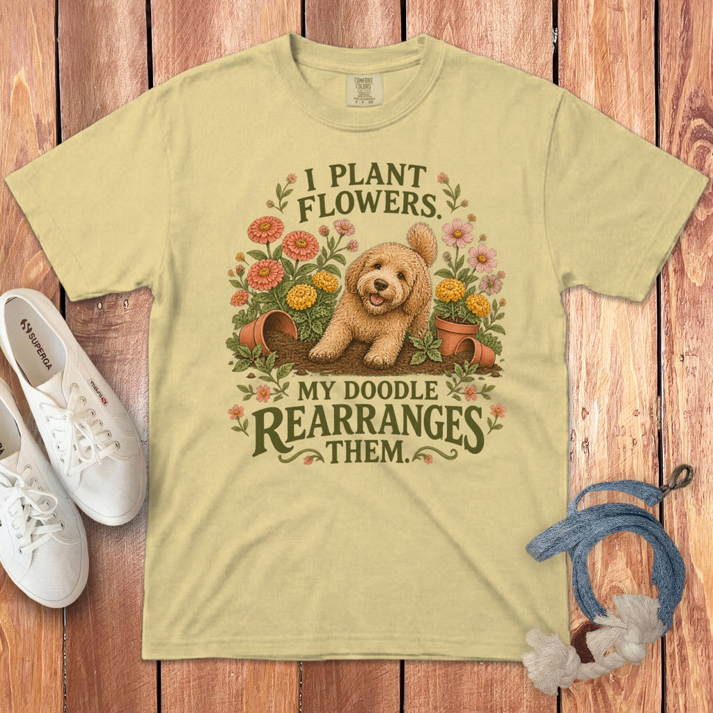 Light yellow t-shirt with a cartoon goldendoodle graphic, flowers, and the text I PLANT FLOWERS. MY DOODLE REARRANGES THEM.