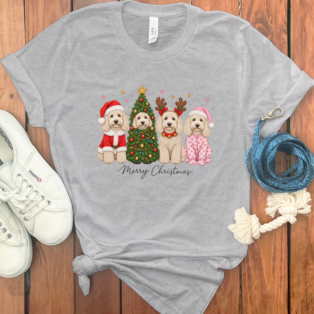 A gray t-shirt with a graphic of four doodle dogs celebrating Christmas, surrounded by sneakers and accessories on a wooden background.