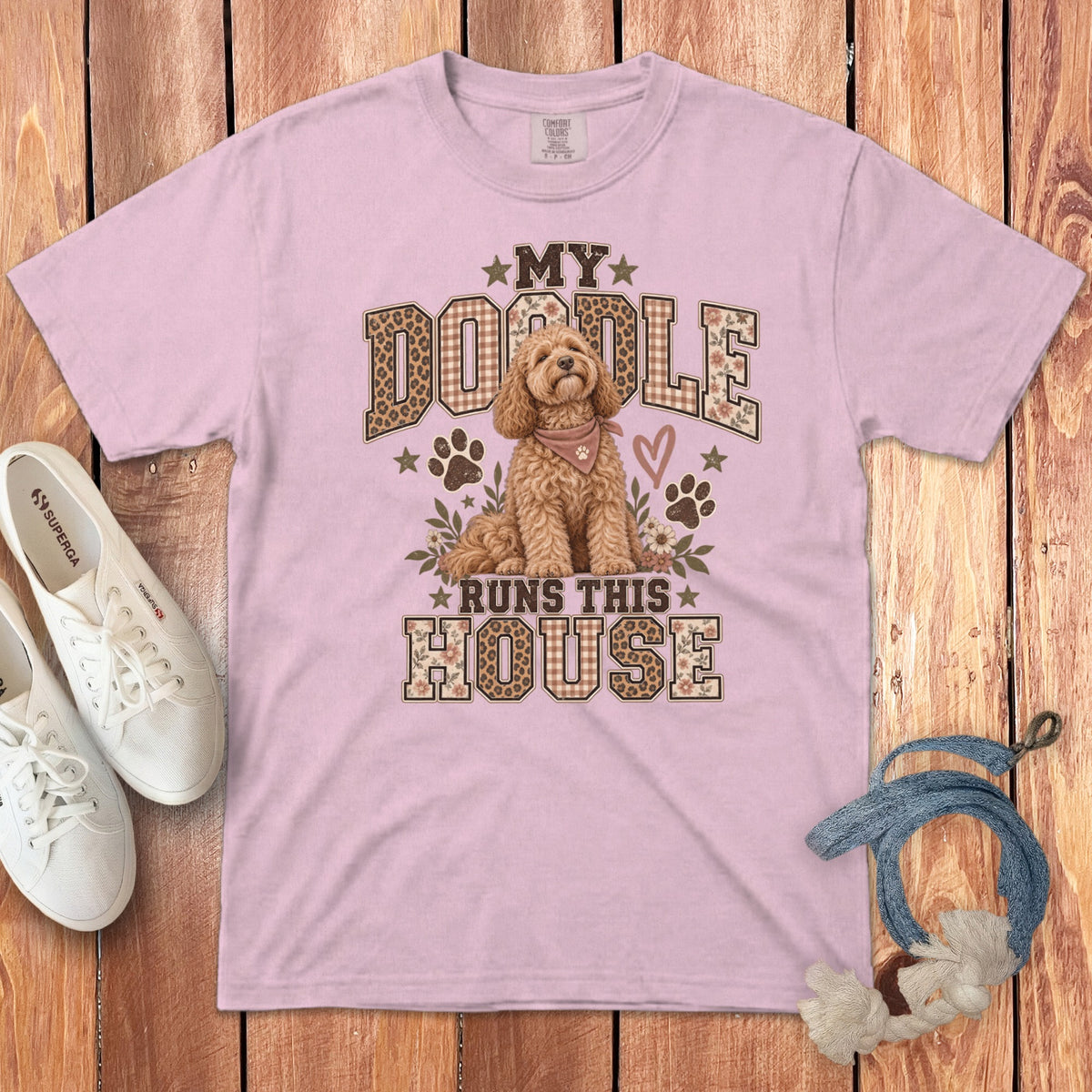 A pink t-shirt with a graphic of a golden doodle and the text My Doodle Runs This House, accompanied by white sneakers and a blue leash on a wooden background.
